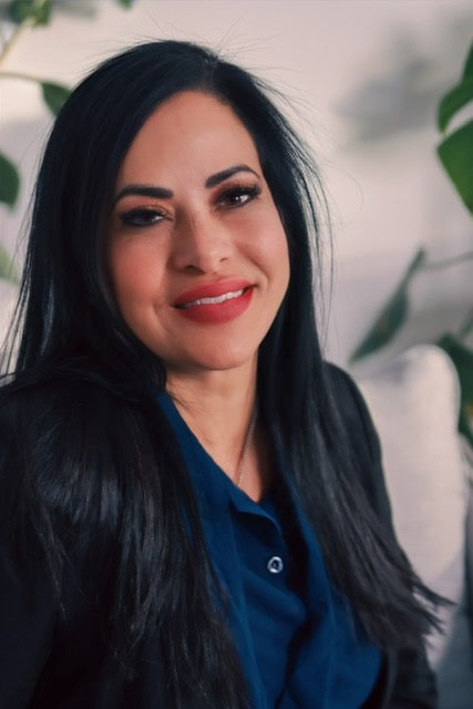 Smiling woman with long dark hair wearing a navy blouse and blazer, seated indoors with soft lighting and plants in the background.